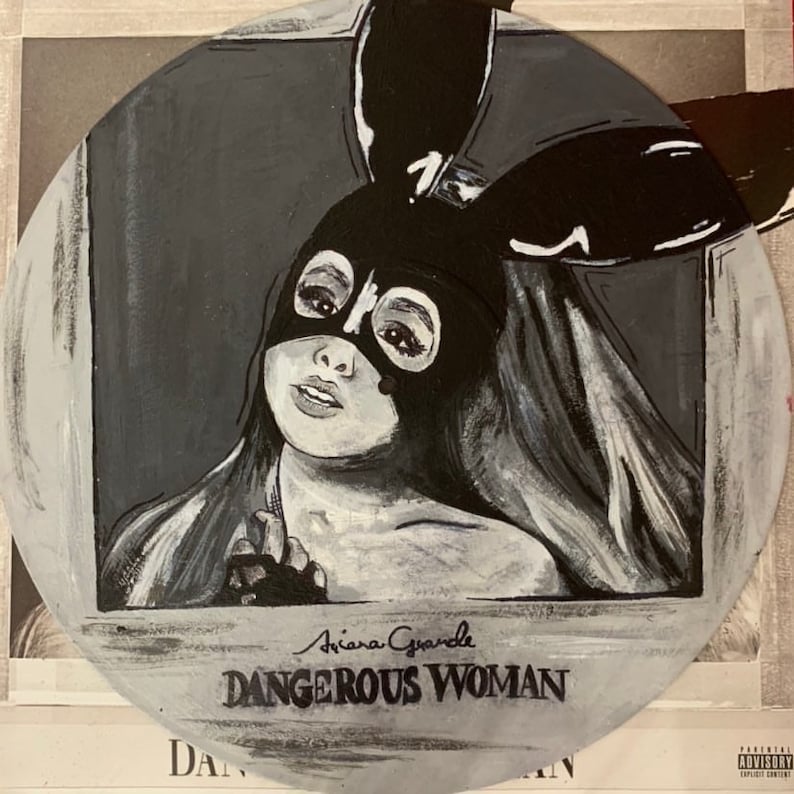 May include: A circular painted vinyl record featuring a black and white portrait of a person wearing a black bunny ear mask. The text Ariana Grande and DANGEROUS WOMAN is painted in black at the bottom of the circular surface against a grey background.