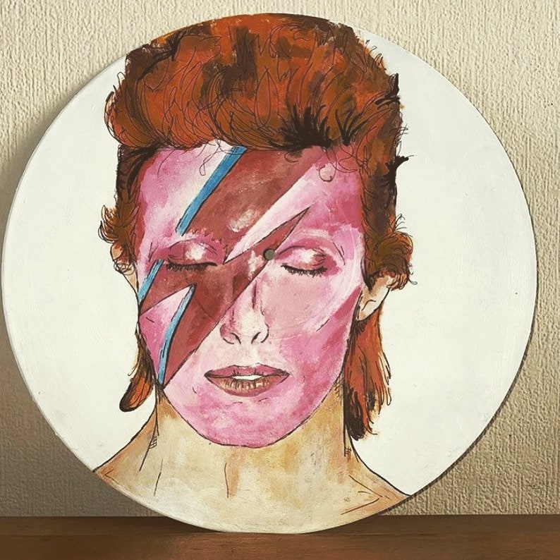 May include: A circular wall art piece featuring a hand-painted portrait of a person with reddish-brown hair and a pink face. A prominent red and blue lightning bolt graphic is painted across the eyes. The artwork is set on a white circular background.