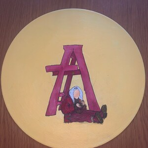 May include: A circular vinyl record painted with a solid yellow background. In the center, a large magenta letter A stands over a figure with light blue hair wearing a dark red hooded jacket and pants, sitting on the ground.