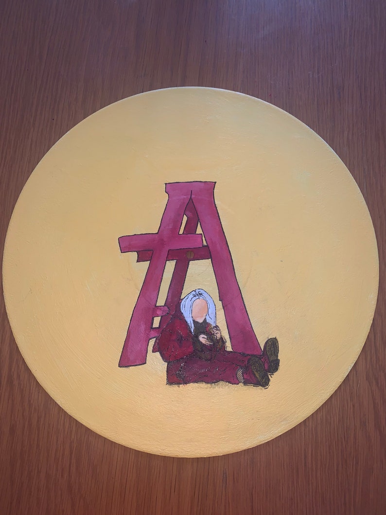 May include: A circular vinyl record painted with a solid yellow background. In the center, a large magenta letter A stands over a figure with light blue hair wearing a dark red hooded jacket and pants, sitting on the ground.
