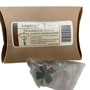 May include: A Longaberger Shamrock Knob in a clear plastic bag, positioned in front of a brown cardboard pillow box. The box features a white label with the text Longaberger, Shamrock Knob, item number 23282, and instructions for removal.
