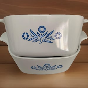 May include: Two white rectangular casserole dishes stacked on top of each other. Each dish has a blue floral design and two handles. The floral design includes flowers and leaves.
