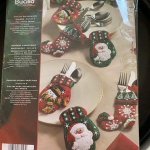 May include: A package of Bucilla felt home decor, including six Christmas-themed silverware holders. The holders are shaped like mittens and stockings, featuring Santa Claus, snowmen, and snowflake designs. The package includes instructions and materials.