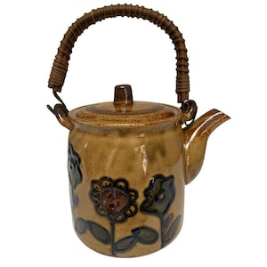 May include: A tan ceramic teapot with a woven handle and lid. The teapot features embossed floral designs in shades of brown, green, and blue. The spout and handle are brown.