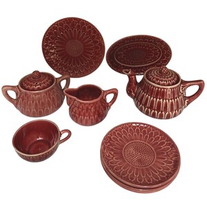 May include: A maroon ceramic tea set featuring a teapot, creamer, teacup, and plates. The plates have a sunflower design, and the teapot and creamer have a textured pattern. The set is designed for serving tea.