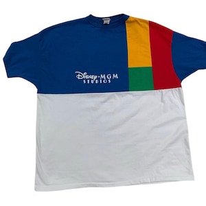 May include: A blue, white, red, green, and yellow color block t-shirt with the text "Disney MGM Studios" in white. The shirt has a crew neck and short sleeves. The top half of the shirt is blue, with a white section below.