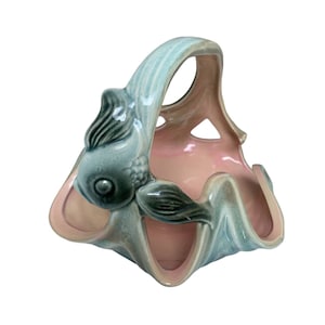 May include: A ceramic basket with a fish design. The basket is light blue on the outside and pink on the inside. The handle is arched, and a fish is sculpted on the side. The basket has a wavy, open design.