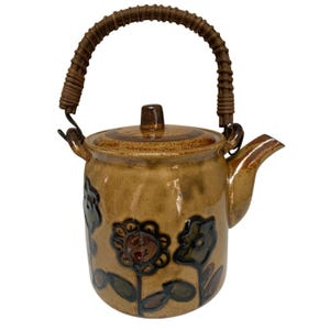 May include: A tan ceramic teapot with a glossy finish, featuring dark brown hand-painted floral motifs on the body. The teapot includes a matching lid and a curved, textured brown woven handle attached to two metal loops on the sides of the vessel.