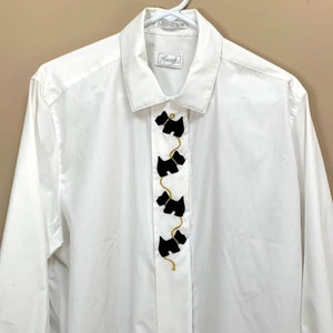 May include: White button-down shirt with long sleeves. The shirt features a vertical line of four black Scottie dog appliqués connected by gold-colored thread. The shirt has a collar and a label that reads "Foxcraft".