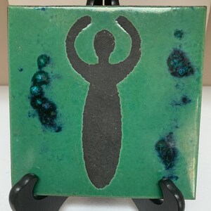 May include: A square, green ceramic tile with a black silhouette of a person with raised arms. The tile has dark blue and black accents and is displayed on a black easel.