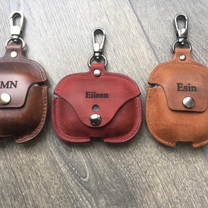 May include: Three personalized leather AirPod cases with a carabiner clip. The cases are brown, red, and tan. The names "JMN", "Eileen", and "Esin" are engraved on the cases.