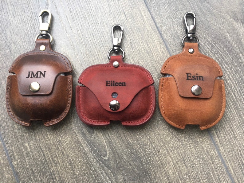 May include: Three personalized leather AirPod cases with a carabiner clip. The cases are brown, red, and tan. The names "JMN", "Eileen", and "Esin" are engraved on the cases.