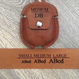 May include: Brown leather Airpods case with a silver snap closure. The case has the words "MEDIUM" and "DB" engraved on it. The case is attached to a silver keychain. A brown leather strap with the words "SMALL MEDIUM LARGE" and "ABcd ABcd ABcd" engraved on it is shown below the case.