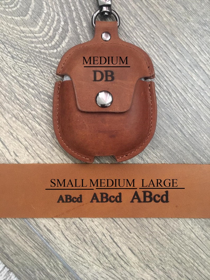 May include: Brown leather Airpods case with a silver snap closure. The case has the words "MEDIUM" and "DB" engraved on it. The case is attached to a silver keychain. A brown leather strap with the words "SMALL MEDIUM LARGE" and "ABcd ABcd ABcd" engraved on it is shown below the case.