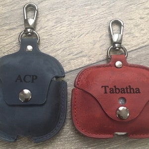 May include: Two personalized leather Airpod cases with silver clasps. The case on the left is dark blue with the name "ACP" engraved on it. The case on the right is burgundy with the name "Tabatha" engraved on it.