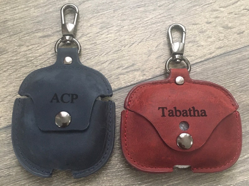 May include: Two personalized leather Airpod cases with silver clasps. The case on the left is dark blue with the name "ACP" engraved on it. The case on the right is burgundy with the name "Tabatha" engraved on it.