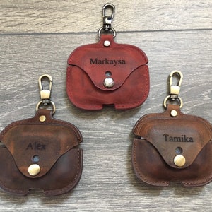 May include: Three personalized leather Airpod cases with a gold-tone metal carabiner clip. The cases are brown, dark brown, and red. The names "Markaysa", "Alex", and "Tamika" are engraved on the cases.