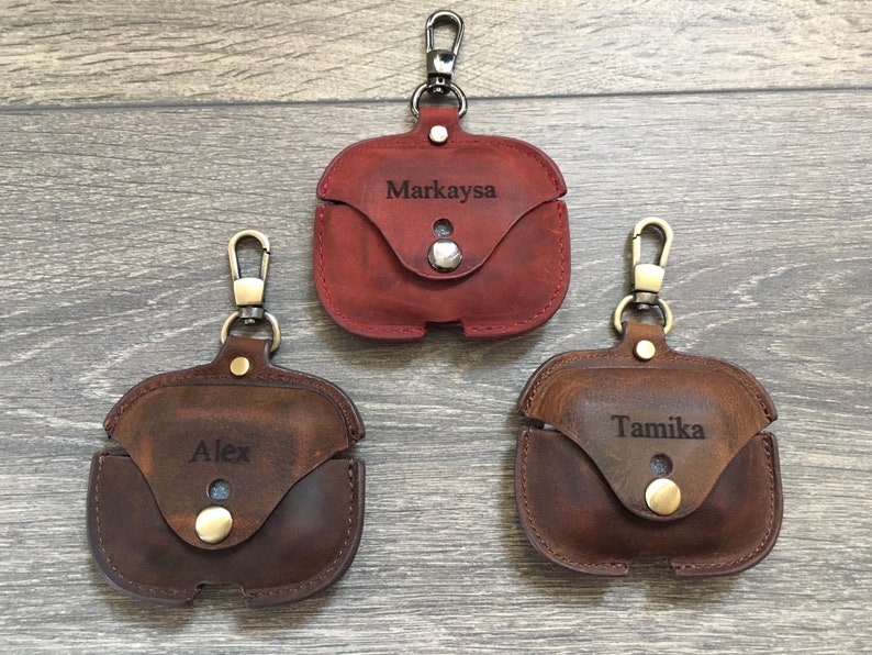May include: Three personalized leather Airpod cases with a gold-tone metal carabiner clip. The cases are brown, dark brown, and red. The names "Markaysa", "Alex", and "Tamika" are engraved on the cases.