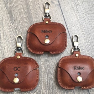 May include: Three brown leather Airpod cases with a metal keychain loop. The cases are personalized with the names "Mikey", "GC", and "Khloe".