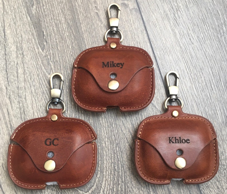 May include: Three brown leather Airpod cases with a metal keychain loop. The cases are personalized with the names "Mikey", "GC", and "Khloe".