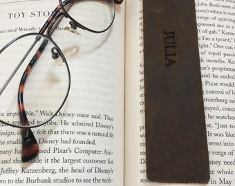 May include: A close-up of a brown leather bookmark with the name "Julia" engraved in gold lettering. The bookmark is resting on an open book with the title "Toy Story" visible.
