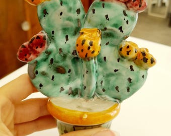 May include: A ceramic cactus figurine featuring a glossy green body with dark speckles, potted in a round yellow base. The cactus has several rounded pads adorned with textured orange and red flower buds attached to the top and side edges.
