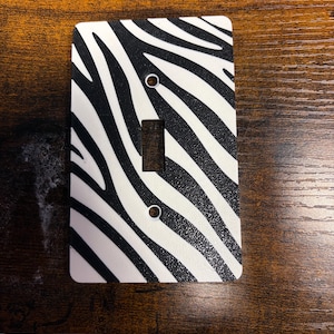 May include: A rectangular light switch cover plate featuring a black and white zebra stripe pattern. The plate has a central rectangular cutout for a toggle switch and two circular screw holes positioned vertically above and below the center.