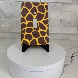 May include: A rectangular light switch wall plate featuring a brown and yellow giraffe print pattern. The plate has a central vertical rectangular cutout for a toggle switch and two circular screw holes positioned at the top and bottom center.