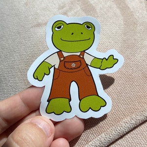 May include: A die-cut sticker featuring a cartoon frog character. The frog is green with a white shirt, brown overalls, and a small flower detail. The sticker has a white border.