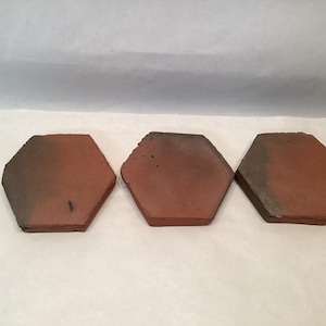May include: Three hexagonal terracotta tiles. Each tile has a reddish-brown hue with darker edges. The tiles are arranged in a row, showcasing their shape and color variations. These tiles are suitable for various home decor projects.