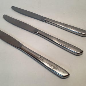 May include: Three stainless steel dinner knives with a floral pattern etched on the handles. The blades are smooth and the handles have a rounded shape. The knives are arranged diagonally on a white surface.