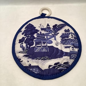 May include: Round blue and white pot holder with a classic Asian-inspired landscape design. The pot holder has a dark blue trim and a white loop for hanging. The design features pagodas, trees, and water elements.