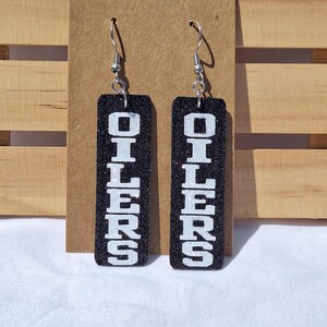May include: A pair of rectangular dangle earrings featuring a black glitter finish and the word OILERS printed in bold white block letters. The earrings are attached to silver-toned metal hooks and hang vertically from a brown cardboard display card.
