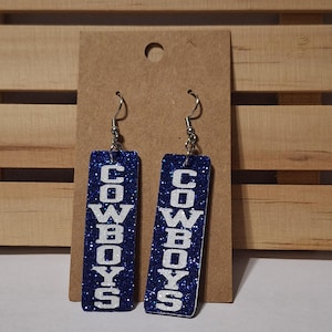 Pode incluir: Um par de brincos pendurados com fundo azul brilhante e a palavra "COWBOYS" em letras brancas. Os brincos são retangulares e pendem de ganchos prateados. Os brincos são exibidos em um suporte de papelão marrom.