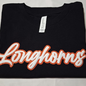 Puede incluir: Una camiseta negra de manga corta con la palabra Longhorns en una fuente cursiva blanca con un contorno grueso de purpurina naranja texturizada. La camiseta tiene una etiqueta blanca en el cuello con la marca Bella Canvas en negro.