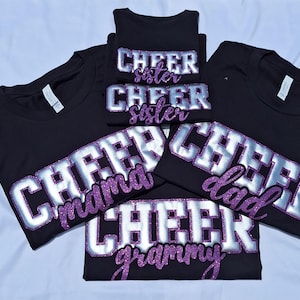 K&ouml;nnte beinhalten: Eine Sammlung von f&uuml;nf schwarzen T-Shirts mit dem Wort CHEER in gro&szlig;en silbernen Blockbuchstaben mit violetten Glitzerumrandungen. Unter dem Wort CHEER zeigt jedes T-Shirt einen spezifischen Titel in violetter Glitzerschrift: sister, mama, dad, grammy.
