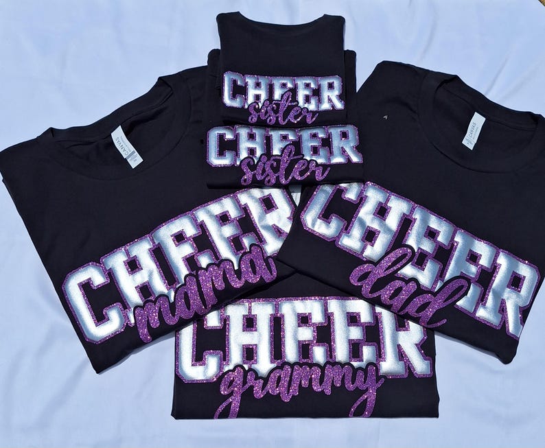 K&ouml;nnte beinhalten: Eine Sammlung von f&uuml;nf schwarzen T-Shirts mit dem Wort CHEER in gro&szlig;en silbernen Blockbuchstaben mit violetten Glitzerumrandungen. Unter dem Wort CHEER zeigt jedes T-Shirt einen spezifischen Titel in violetter Glitzerschrift: sister, mama, dad, grammy.