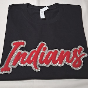 Könnte beinhalten: Ein schwarzes T-Shirt mit kurzen Ärmeln, das das Wort Indians in einer roten Schreibschrift zeigt. Die rote Schrift ist mit einem silbernen Glitzerrand versehen. Ein weißes Markenetikett ist innen am Rundhalsausschnitt auf dem Stoff sichtbar.