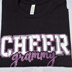 K&ouml;nnte beinhalten: Ein schwarzes Kurzarm-T-Shirt mit dem Wort CHEER in gro&szlig;en silbernen Blockbuchstaben mit violettem Glitzerumriss und dem Wort grammy in violetter Glitzerschrift darunter. Das T-Shirt hat ein wei&szlig;es Etikett am Halsausschnitt.