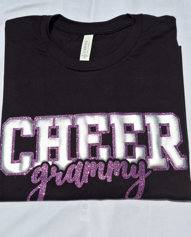 K&ouml;nnte beinhalten: Ein schwarzes Kurzarm-T-Shirt mit dem Wort CHEER in gro&szlig;en silbernen Blockbuchstaben mit violettem Glitzerumriss und dem Wort grammy in violetter Glitzerschrift darunter. Das T-Shirt hat ein wei&szlig;es Etikett am Halsausschnitt.