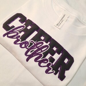 K&ouml;nnte beinhalten: Ein wei&szlig;es T-Shirt mit dem Wort CHEER in schwarzen Blockbuchstaben und dem Wort brother in violetter Glitzerschrift. Das Shirt hat ein wei&szlig;es Nackenetikett mit dem in schwarzer Tinte gedruckten Text BELLA+CANVAS AIRLUME JERSEY.
