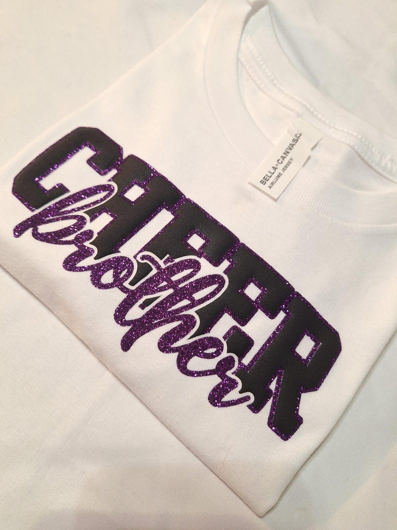 K&ouml;nnte beinhalten: Ein wei&szlig;es T-Shirt mit dem Wort CHEER in schwarzen Blockbuchstaben und dem Wort brother in violetter Glitzerschrift. Das Shirt hat ein wei&szlig;es Nackenetikett mit dem in schwarzer Tinte gedruckten Text BELLA+CANVAS AIRLUME JERSEY.
