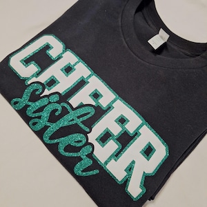 K&ouml;nnte beinhalten: Ein schwarzes T-Shirt mit kurzen &Auml;rmeln und dem Schriftzug CHEER SISTER auf der Vorderseite. Das Wort CHEER besteht aus gro&szlig;en wei&szlig;en Blockbuchstaben mit einem gr&uuml;nen Glitzerumriss, das Wort SISTER ist in gr&uuml;ner Glitzerschrift gehalten.