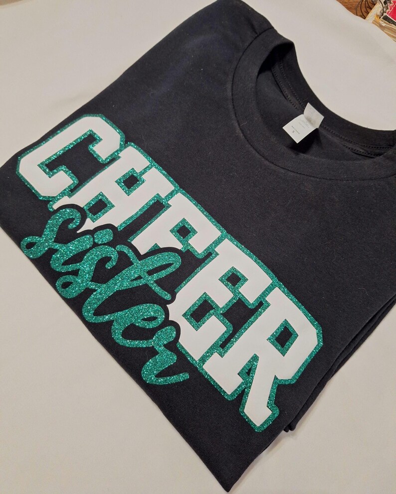 K&ouml;nnte beinhalten: Ein schwarzes T-Shirt mit kurzen &Auml;rmeln und dem Schriftzug CHEER SISTER auf der Vorderseite. Das Wort CHEER besteht aus gro&szlig;en wei&szlig;en Blockbuchstaben mit einem gr&uuml;nen Glitzerumriss, das Wort SISTER ist in gr&uuml;ner Glitzerschrift gehalten.