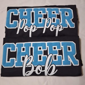 K&ouml;nnte beinhalten: Zwei schwarze T-Shirts mit dem Wort CHEER in gro&szlig;en, hellblauen Blockbuchstaben mit wei&szlig;em Glitzerumriss. Das obere T-Shirt zeigt den Text Pop Pop in wei&szlig;er Schreibschrift, das untere T-Shirt zeigt den Text Bob in wei&szlig;er Schreibschrift.