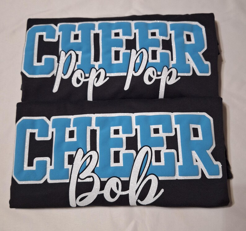 K&ouml;nnte beinhalten: Zwei schwarze T-Shirts mit dem Wort CHEER in gro&szlig;en, hellblauen Blockbuchstaben mit wei&szlig;em Glitzerumriss. Das obere T-Shirt zeigt den Text Pop Pop in wei&szlig;er Schreibschrift, das untere T-Shirt zeigt den Text Bob in wei&szlig;er Schreibschrift.