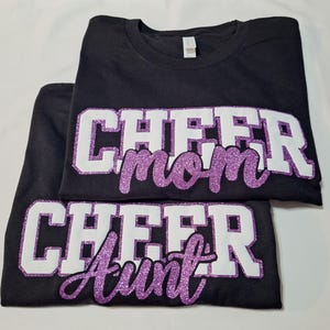 K&ouml;nnte beinhalten: Zwei schwarze T-Shirts mit wei&szlig;er Blockschrift und lilafarbenem Glitzer-Schriftzug. Das obere Shirt zeigt den Text CHEER mom, das untere Shirt zeigt den Text CHEER Aunt. Beide Shirts haben einen Rundhalsausschnitt und kurze &Auml;rmel.