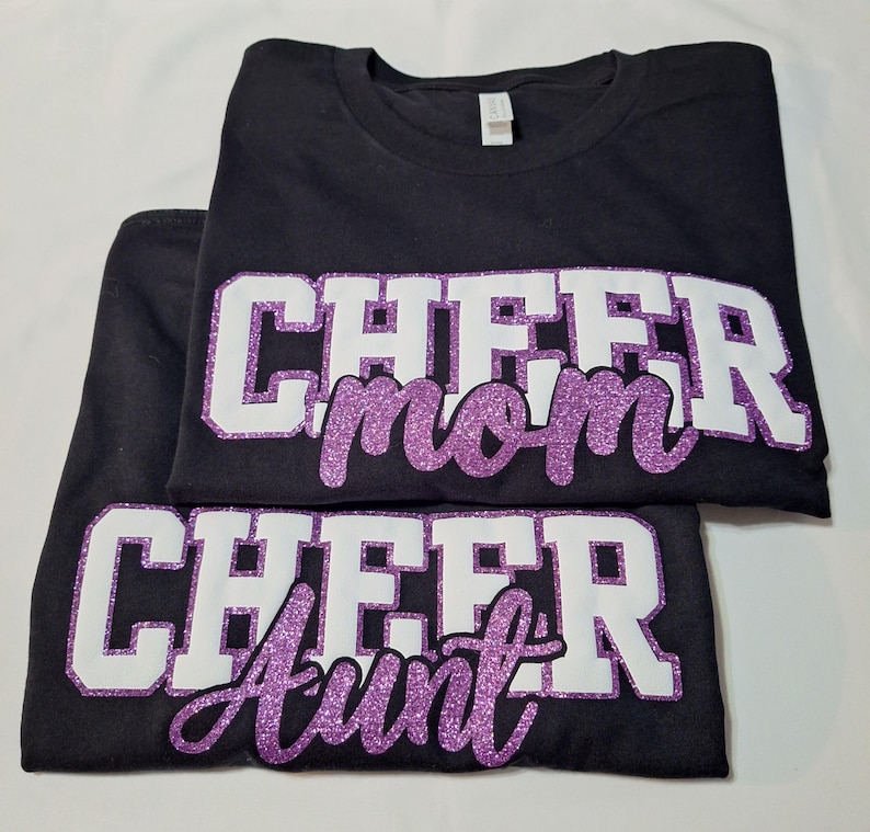 K&ouml;nnte beinhalten: Zwei schwarze T-Shirts mit wei&szlig;er Blockschrift und lilafarbenem Glitzer-Schriftzug. Das obere Shirt zeigt den Text CHEER mom, das untere Shirt zeigt den Text CHEER Aunt. Beide Shirts haben einen Rundhalsausschnitt und kurze &Auml;rmel.