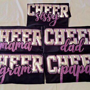 K&ouml;nnte beinhalten: F&uuml;nf schwarze T-Shirts mit dem Wort CHEER in gro&szlig;en silbernen Blockbuchstaben mit violetten Glitzerumrandungen. Unter dem Wort CHEER zeigt jedes T-Shirt ein einzigartiges Wort in violetter Glitzerschrift: sissy, mama, dad, gram oder papa.