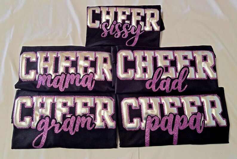 K&ouml;nnte beinhalten: F&uuml;nf schwarze T-Shirts mit dem Wort CHEER in gro&szlig;en silbernen Blockbuchstaben mit violetten Glitzerumrandungen. Unter dem Wort CHEER zeigt jedes T-Shirt ein einzigartiges Wort in violetter Glitzerschrift: sissy, mama, dad, gram oder papa.